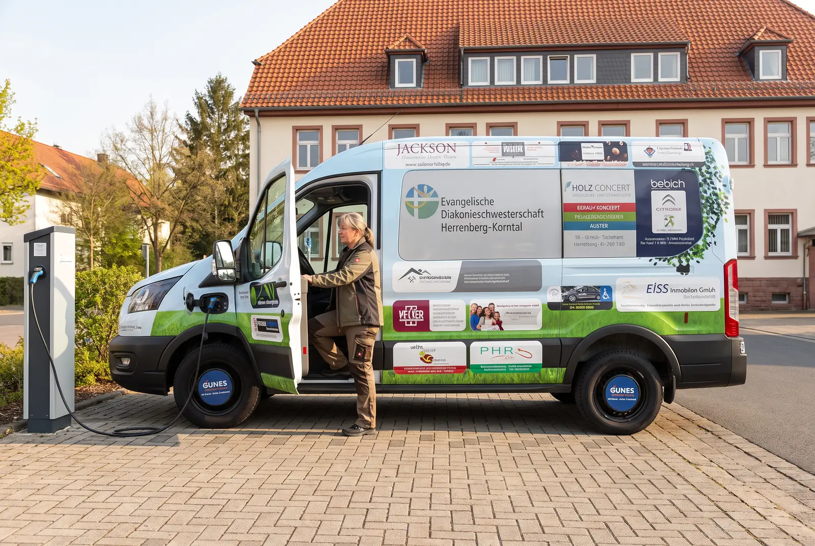 Ein kompakter Hybrid-Kleinwagen in heller Lackierung steht vor einer sozialen Einrichtung. Auf den Türen sind dezent gestaltete Logos lokaler Sponsoren foliert. Tageslicht, leicht diesig. Ruhiges, alltägliches Stadt-Setting.